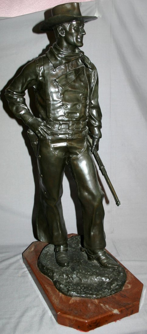 BRONZE SCULPTURE, 29" H. X 10" W., "JOHN WAYNE": BRONZE SCULPTURE, 29" H. X 10" W., "JOHN WAYNE":depicts a standing John Wayne in western garb, a holstered pistol and holding a Winchester rifle, dark green patina; surmounted on a brown marble octago