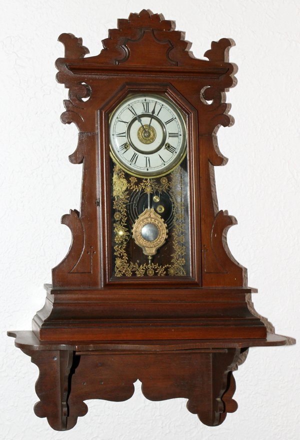 EASTLAKE WALNUT CLOCK WITH SHELF, C1890, H 32": EASTLAKE WALNUT CLOCK WITH SHELF, C1890, H 32", L 19":walnut case with cutout designs on top and front; having a gilt stencil flower and vine design on a glass access door opening to white painted fac