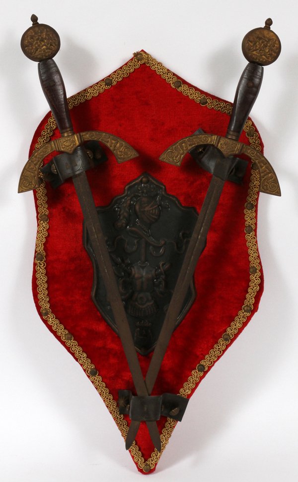 COAT OF ARMS AND CROSSED SWORD WALL PLAQUE 25"H: COAT OF ARMS AND CROSSED SWORD WALL PLAQUE, EARLY 20TH C, H 25", W 14":having a velvet covered shield form plaque trimmed with gold embroidery border with metal crest of Knights helmet and body armor 