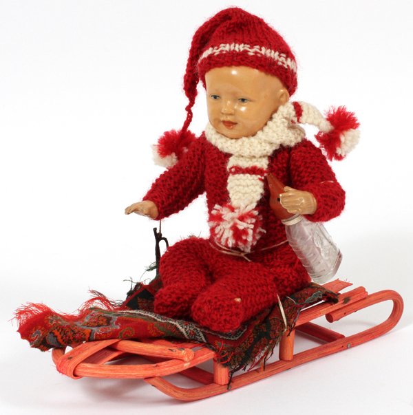 COMPOSITION DOLL SEATED ON SLED C. 1930: COMPOSITION DOLL SEATED ON SLED, C. 1930, H 7", L 8 1/2":Composition doll with painted features and movable limbs, seated on a painted red sled, with a metal horn and glass baby's bottle of the period