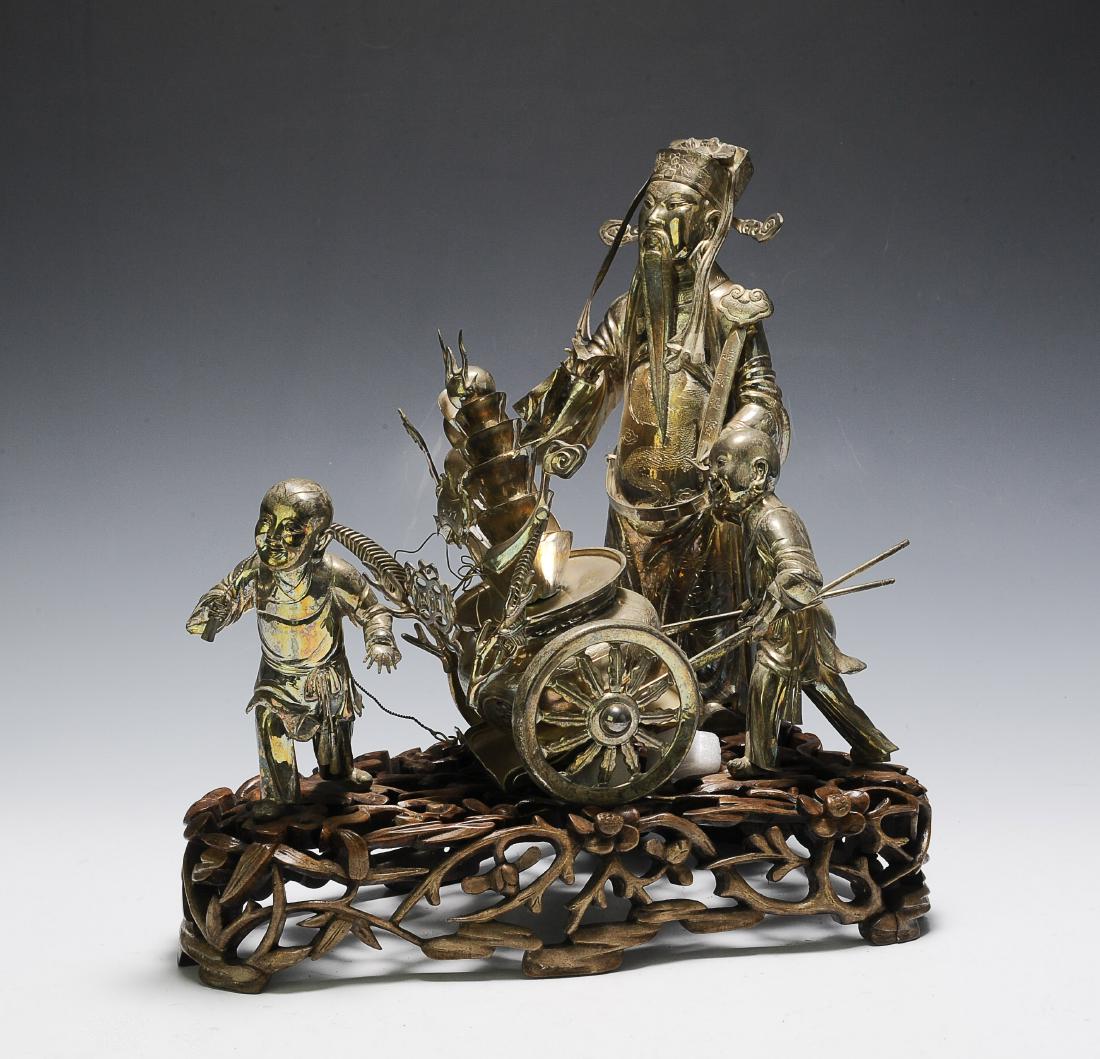 Silver Wealth-Bringing Statue Group, Early 20th C.: A Chinese silver statue group mounted on a wooden base and dating from the early 20th century. The statue group includes Lu holding his ruyi wand and 2 child attendants pulling a chariot of wealth. Th