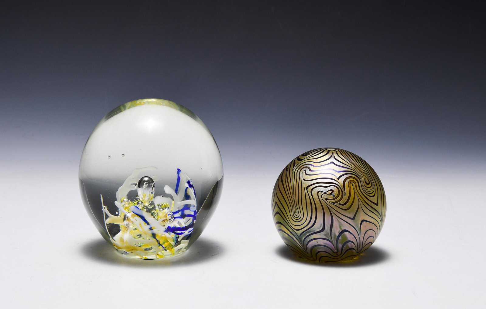 (2) Studio Glass Paperweights, Eickholt & Shriver: (2) studio glass paperweights by Eickholt and Shriver. Shriver paperweight is in yellow, white, and blue in clear glass. Signed Toby '79 on base. Eickholt is in iridescent, feathered gold. Signed Eick