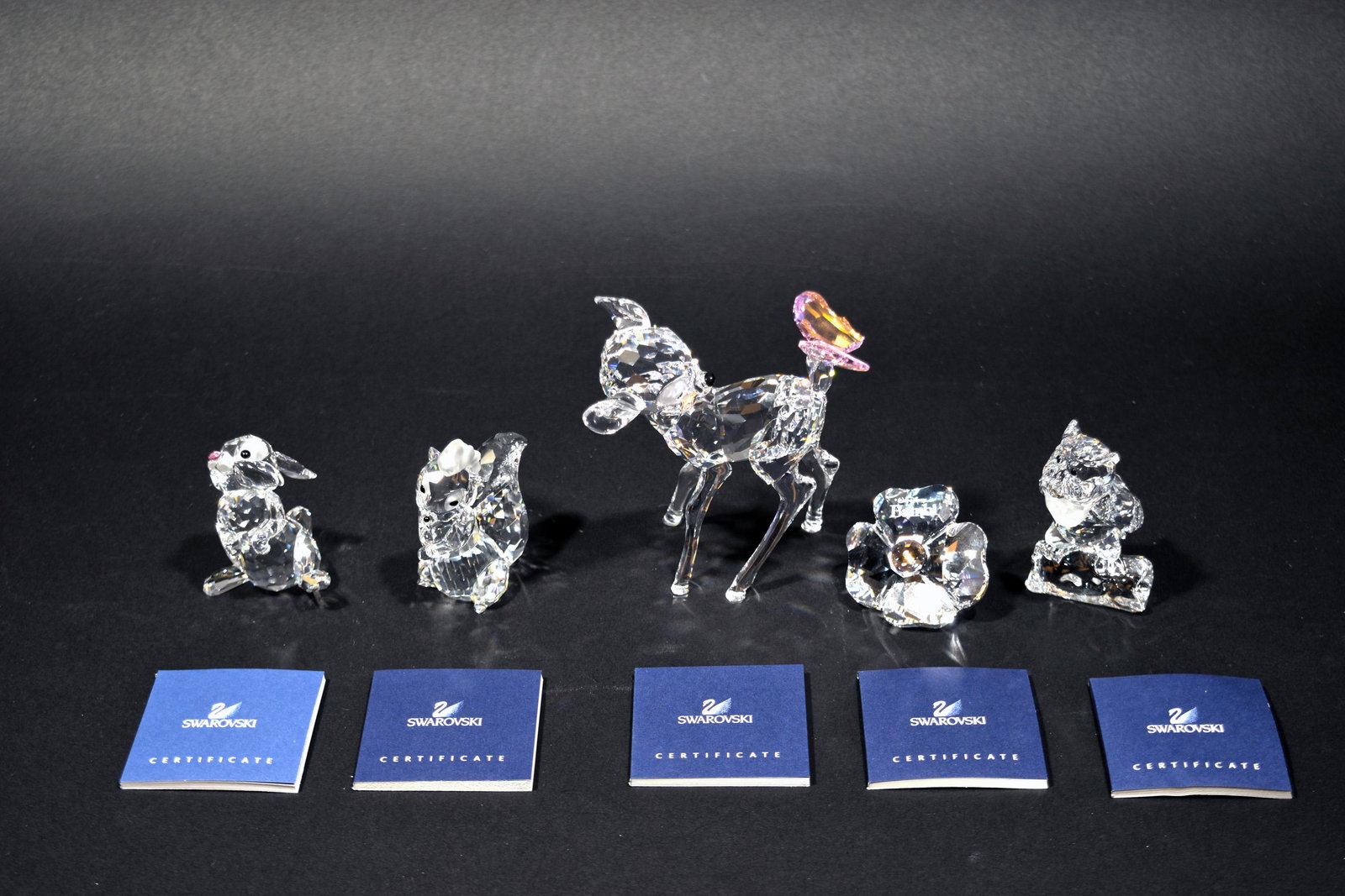 Swarovski, 5 Boxed Bambi Characters: Swarovski (Austrian, founded 1895) crystal. Includes figurines of 'Flower the Skunk,' 'Friend Owl,' 'Thumper' with a pink rose, 'Bambi' with a butterfly on the tail, and a flower-shaped title plaque. 