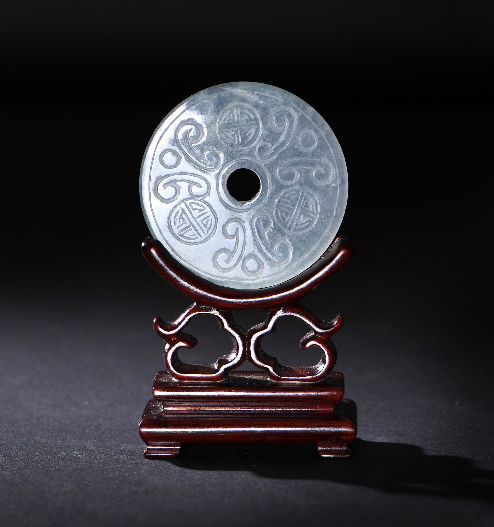 Chinese Jadeite Bi, 19th Century: A Chinese jadeite carved bi dating to the 19th century. One side of the bi features a carving of a flower, while the other side features three Shou medallions and three pairs of ruyi motifs. It is acc