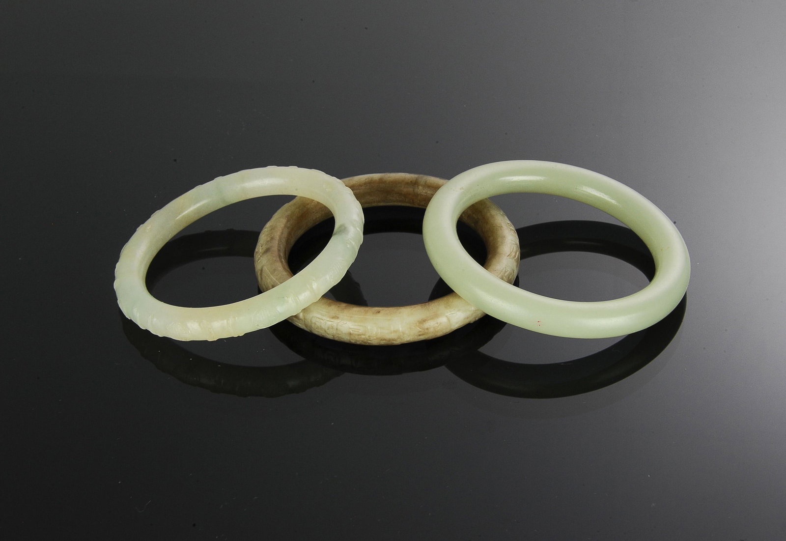 3 Chinese Jade and Jadeite Bangles, Ming- Qing: A group of 3 Chinese jade and jadeite bangles from the Ming and Qing dynasties. Including: a jadeite bangle featuring Shou medallions; a jade bangle; a mutton fat jade carved bangle with 4 groups of 4
