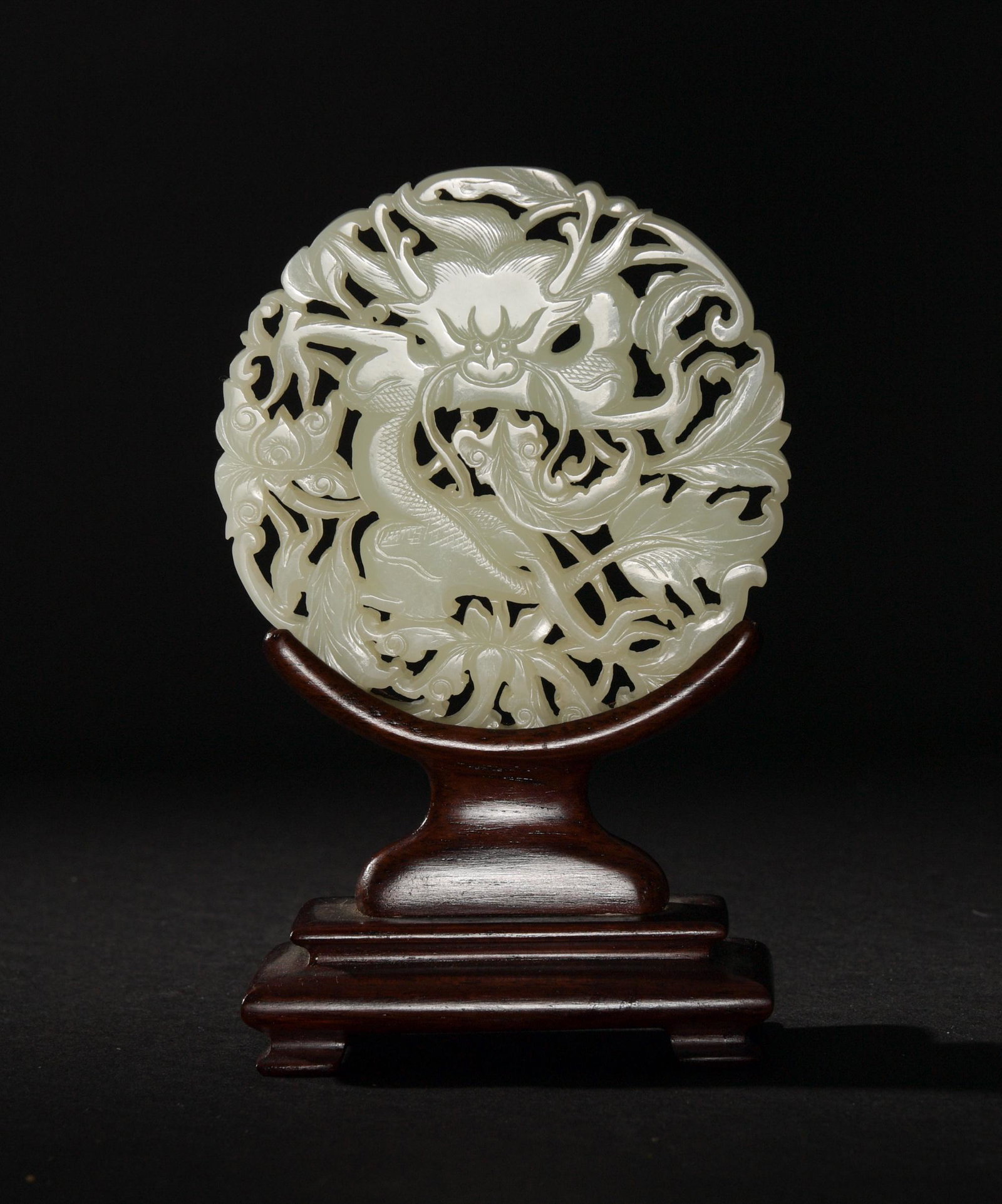 Chinese White Jade Pierced Round Plaque, Ming: A Chinese white jade pierced round plaque dating from the Ming dynasty. The plaque features a dragon among foliage; it comes with a wooden stand. Dimensions: jade- 3 1/4 inches diameter; 8.3 cm diamet