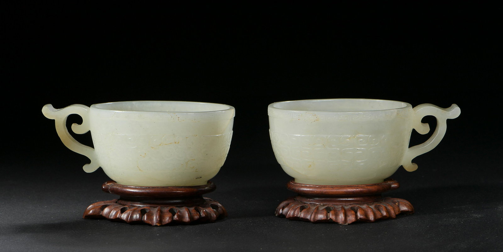 Pair of Chinese Jade Cups, Ming: A pair of small Chinese jade cups dating from the Ming dynasty. The rim of the cups have a Greek key border; they come with wooden stands. Dimensions: 1 1/4 inches tall X 3 inches wide; 3.2 cm tall X 