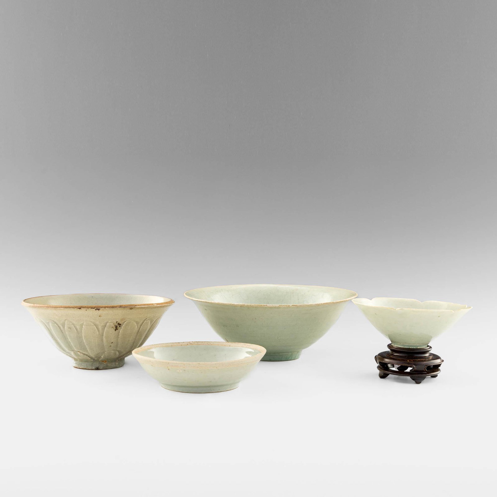 A group of four Chinese yiqing bowls, Song or Yuan dynasty: A group of four Chinese yiqing bowls Song or Yuan dynasty Comprising one bowl with incised fluting to exterior; one small bowl with carved petal-form rim, sold with associated wooden stand; one shallo