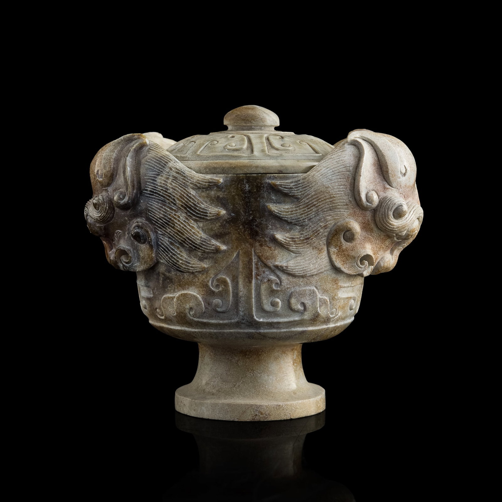 A Chinese carved chicken bone jade lidded cup: A Chinese carved chicken bone jade lidded cup Cup with two finely carved lion heads crowned by flowing manes that cover the cup's body; jade with black and brown inclusions and stylized continuous mot