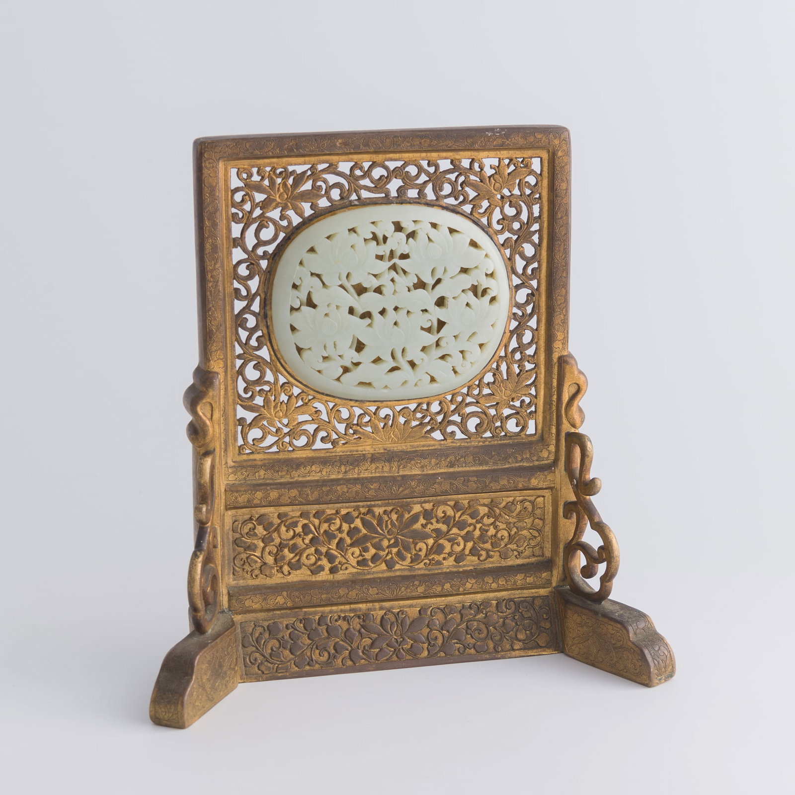 A Chinese gilt-bronze table screen inlaid with jade plaque, late 18th century: A Chinese gilt-bronze table screen inlaid with jade plaque late 18th century Decorated with 'scrolling lotus' motif throughout. Jade: H: 3, W: 3 3/4, D: 1/4 in. (H: 7.6, W: 9.5, D: 0.6 cm.) Overall: H