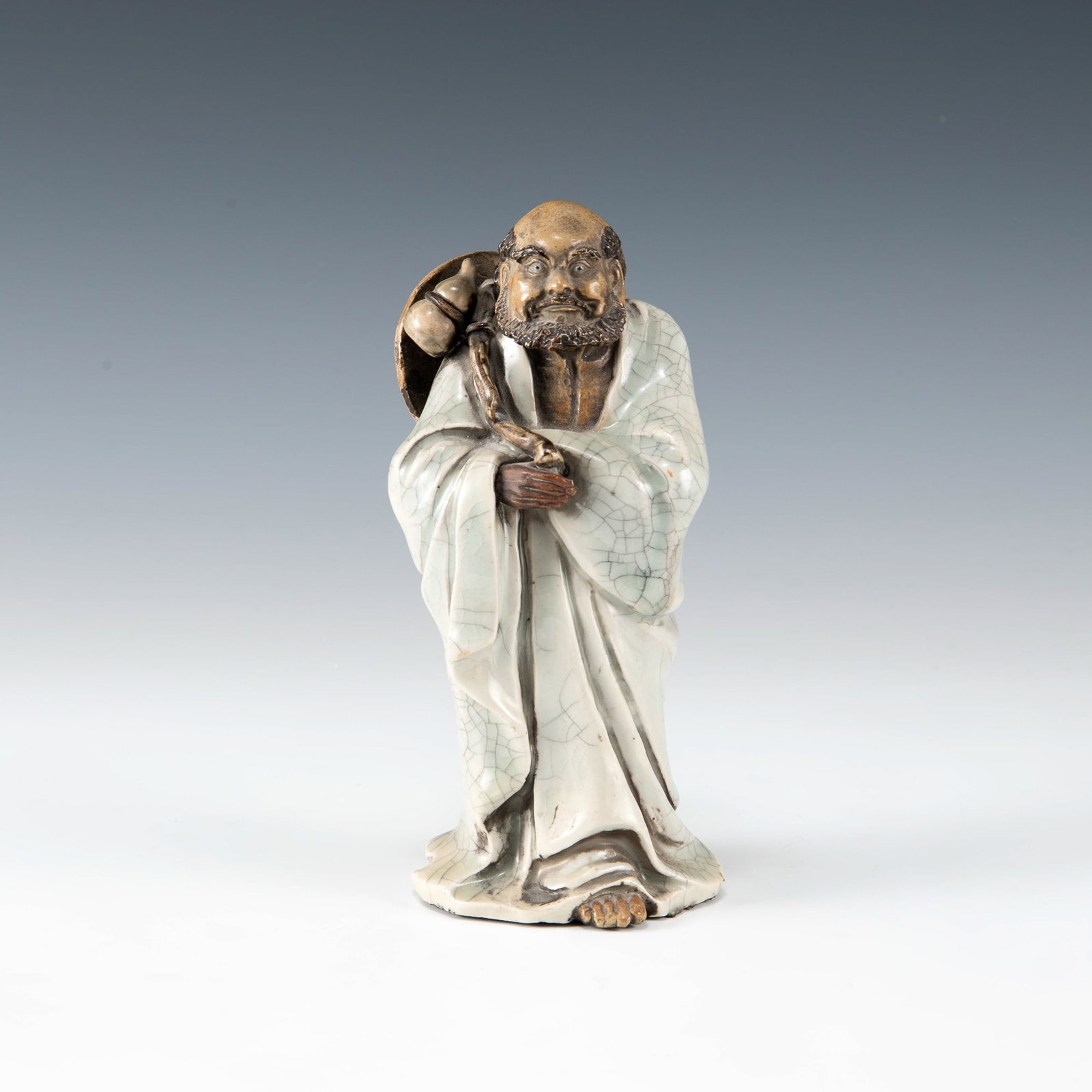 A Chinese Shiwan figure of Damo, Qing dynasty/Republic period: A Chinese Shiwan figure of Damo Qing dynasty/Republic period The standing figure, clothed in a pale blue ge-glazed robe, grips a staff with a hulu gourd, with a straw hat resting on his back. With "Zh
