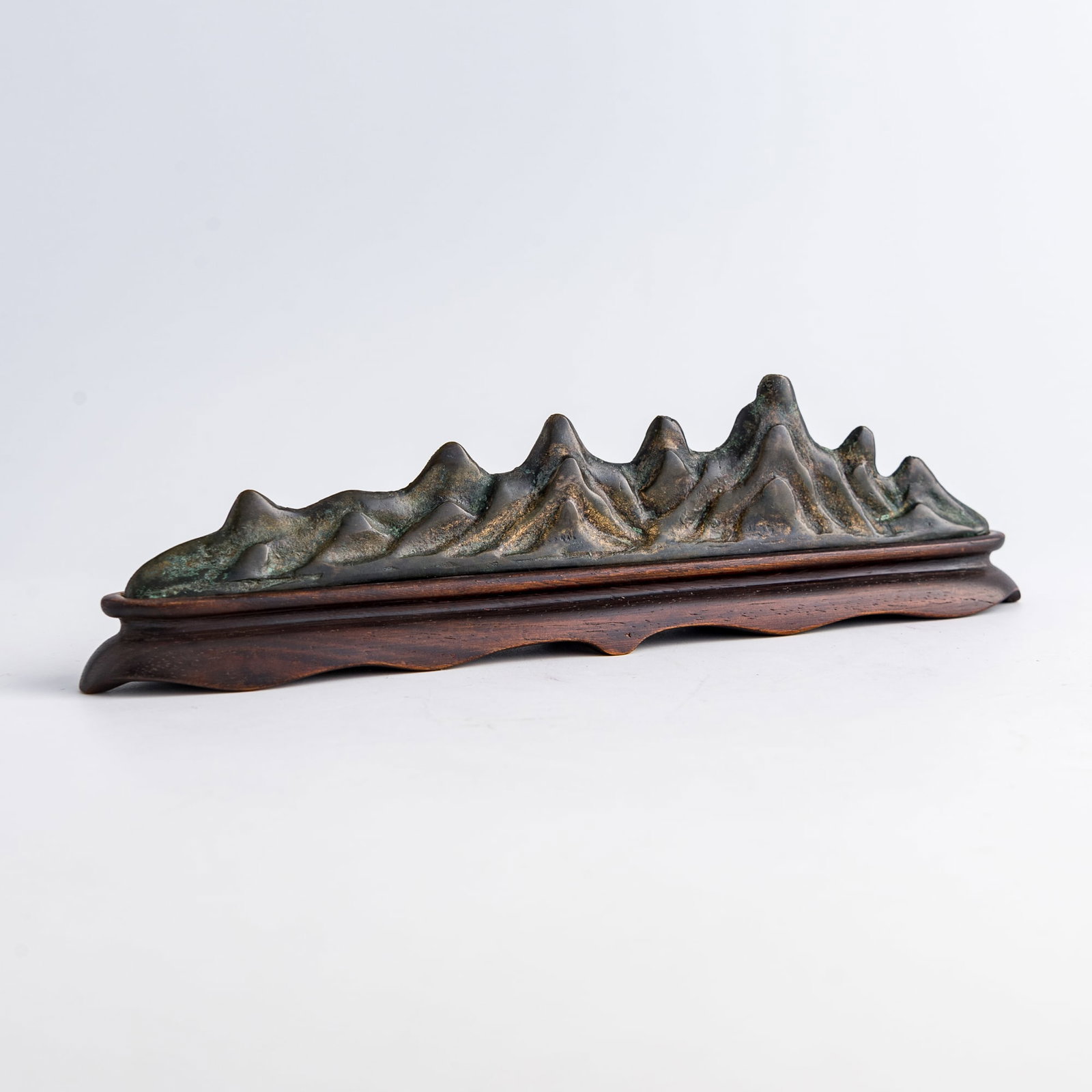 A Chinese bronze mountain-form brush rest, Ming dynasty or earlier: A Chinese bronze mountain-form brush rest Ming dynasty or earlier Sold with original hardwood stand. H: 1 3/4, W: 8 1/2, D: 5/8 in. (H: 4.4, W: 21.6, D: 1.6 cm. (brush rest)) Weight: 301.1 g (brush