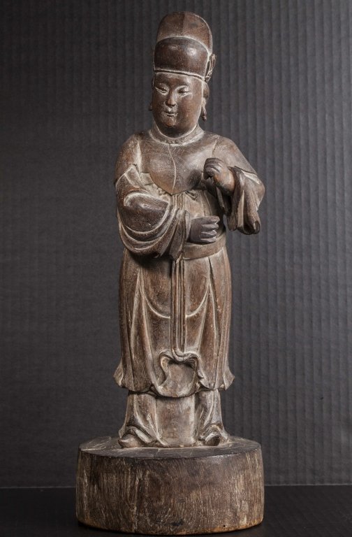 A wood court attendant carving Ming dynasty: A wood carving of a court attendant Ming dynasty the figure is carved as a standing attendant wearing a large headpiece atop his full face his two hands are curved as though he is gripping something a