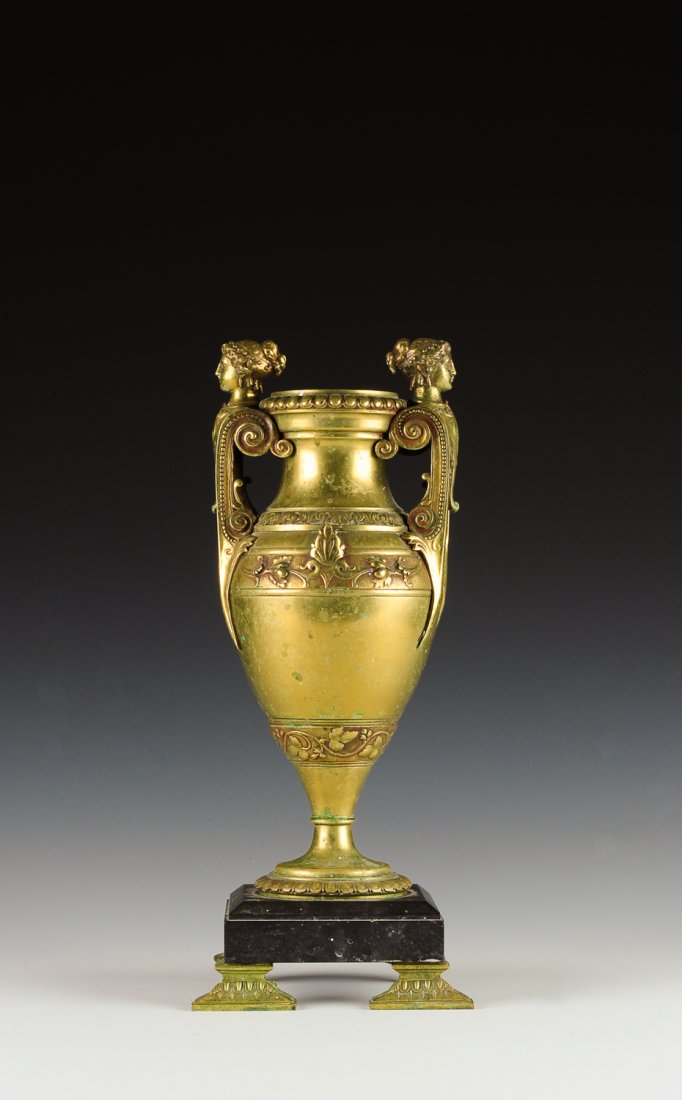 Bronze Neoclassical Amphora w/ Caryatid Handles: Bronze neoclassical style Amphora upon a dark stone base with four brass feet. Two of the feet are round and two are rectangular. The amphora does not have an opening but rather a shallow cup at the m