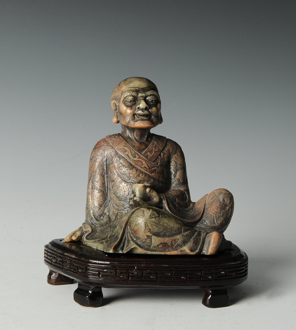 Seated Soapstone Lohan, 20th Century: This seated soapstone figure from the 20th century is of a Lohan in robes holding a cup and can be set in a brown stand. The robes have very fine decorative details. Dimensions are 4 1/2 inches long b