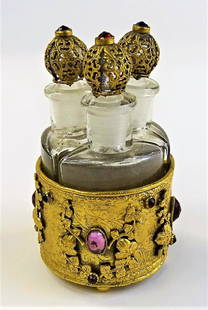 ANTIQUE THREE BOTTLE JEWELED PERFUME CLUSTER: Antique MW & Co. jeweled perfume bottle cluster, containing three triangular shaped bottles with overlayed stoppers. Held in a circular mount set with a variety of colored glass jewels. Overall