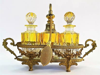 ANTIQUE BACCARAT CRYSTAL & BRASS TRIPLE BOTTLE CADDY: Antique French triple bottle perfume caddy with ornate footed brass mount, decorated with undulating floral garlands, fit with a mirrored plateau. Holding three colored Baccarat crystal bottles, two