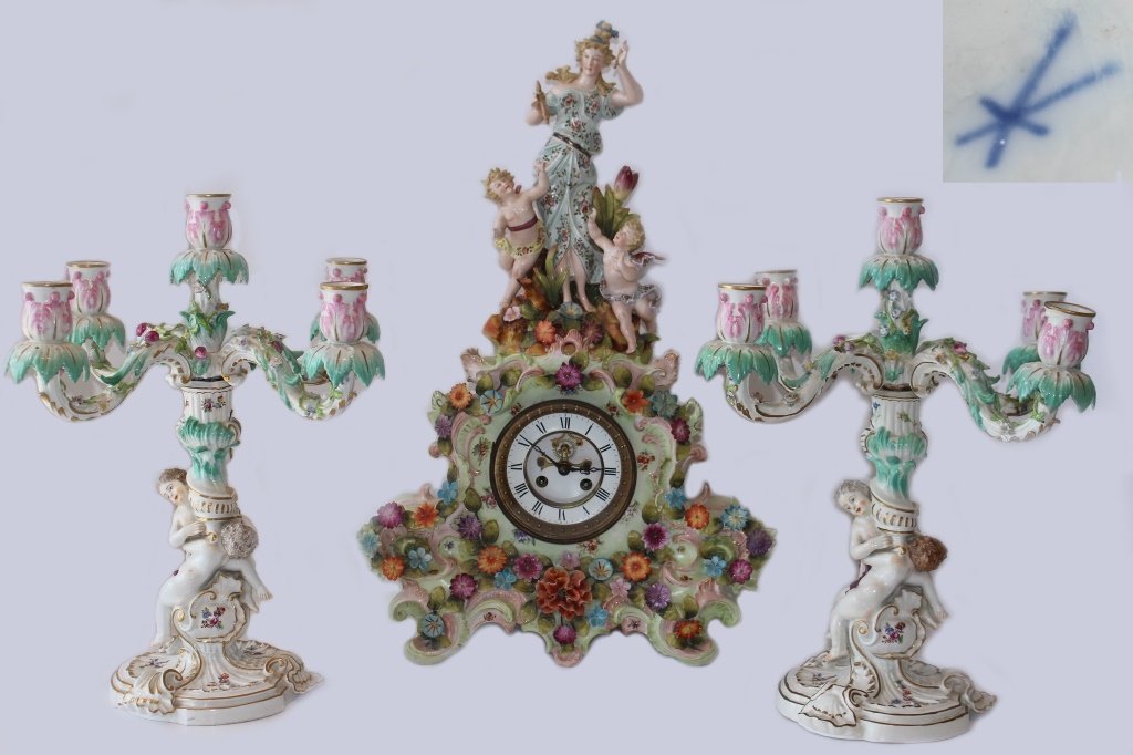 GERMAN ANTIQUE CROSS SWORDS PORCELAIN CLOCK SET: Consisting of a porcelain clock, which measures 22" tall, with woman surrounded by cherubs, sits on a white porcelain clock base with numerous applied flowers. Comes with a pair of matching cherub can
