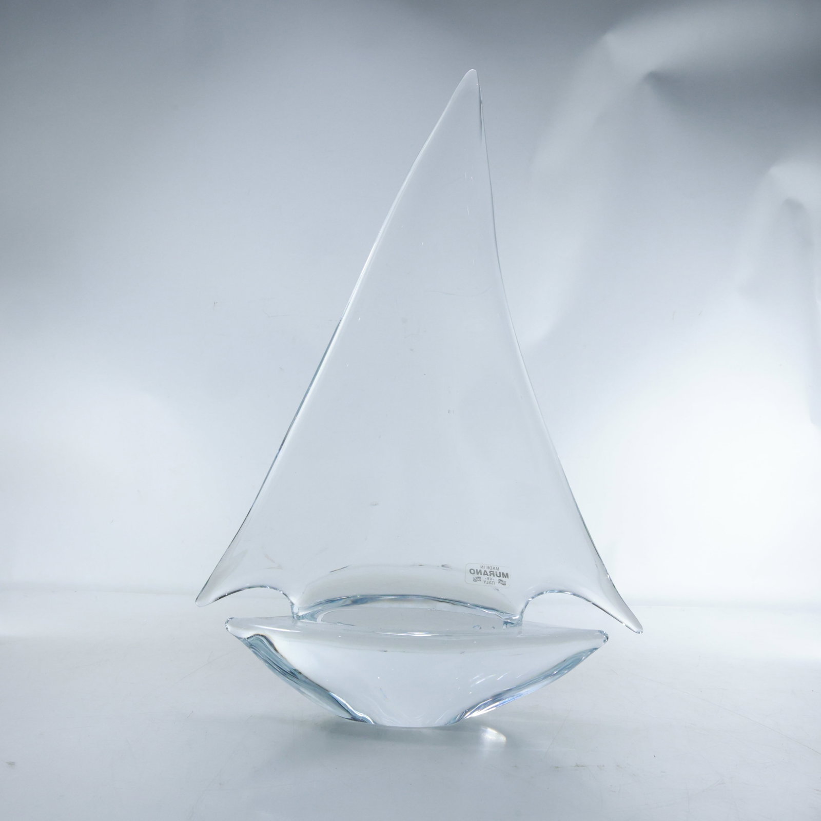 Italian Murano Glass Sailboat - Original Label, 14.5 in. height x 9.5 in. wide: This Murano glass sculpture features a minimalist sailboat design with a sleek, clear glass construction. The sailboat has a simple triangular sail and a smooth, curved hull. The glass is transparent 
