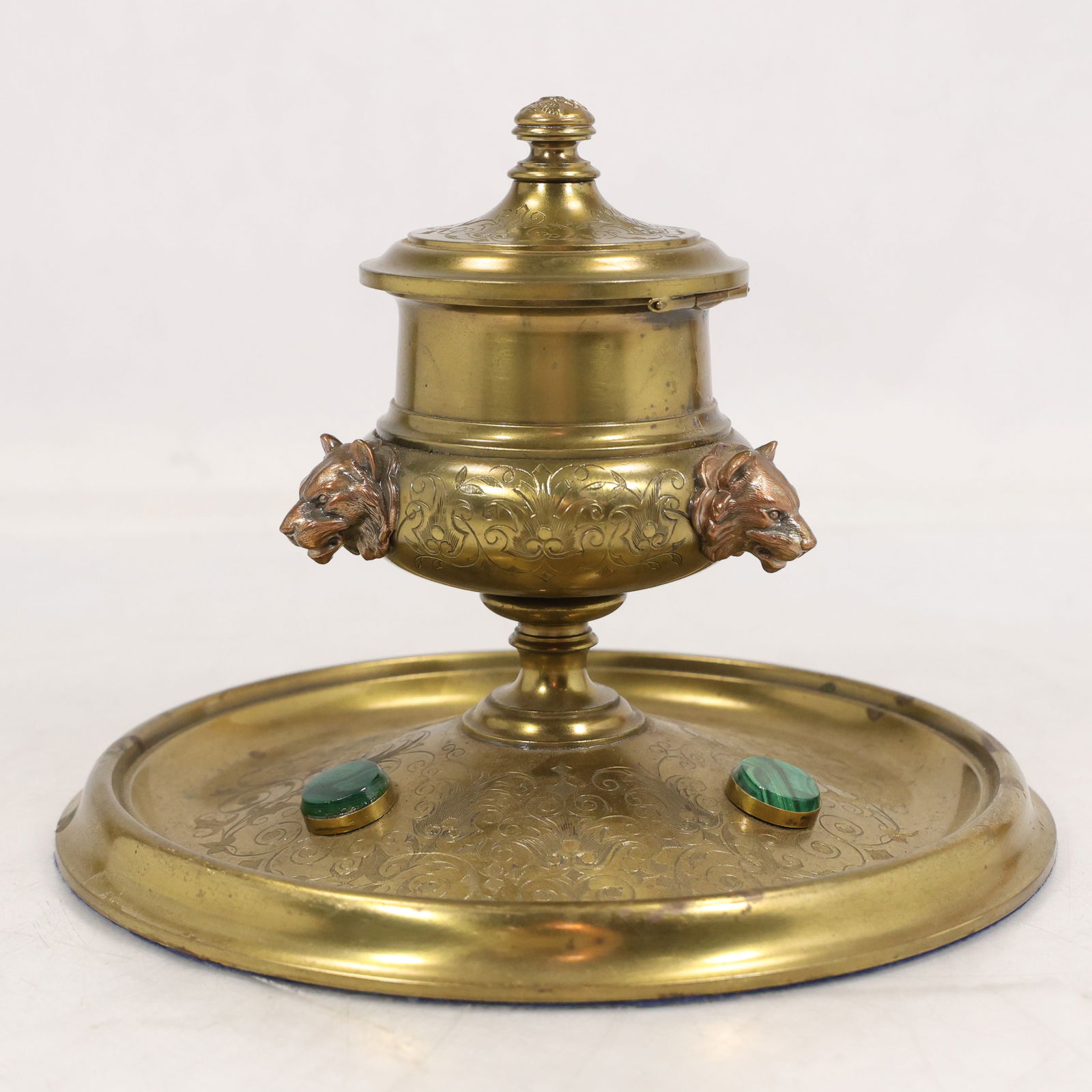 Bronze desk inkwell with wolf head masks and malachite cabochons unmarked: Bronze desk inkwell mounted on a circular engraved base. The piece features a lidded central inkwell with applied wolf head masks on opposite sides and two green malachite cabochons set into the plate