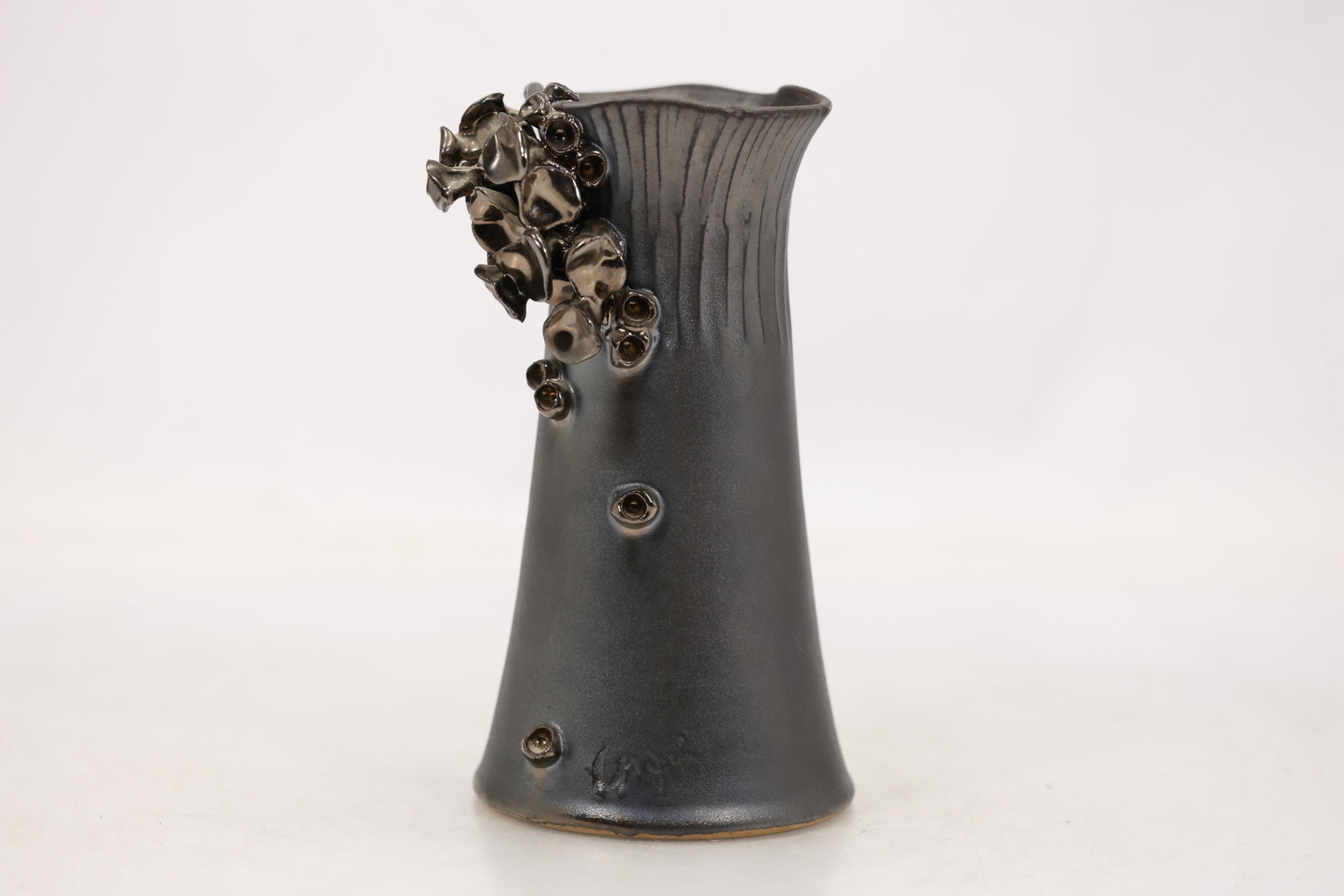 Signed Contemporary Studio Pottery Vase with Applied Metallic Floral Decoration: Tall tapered ceramic vase with a flared irregular rim and vertically incised detailing around the upper body. Finished in a matte charcoal gray glaze with applied sculptural flowers buds and leaves in