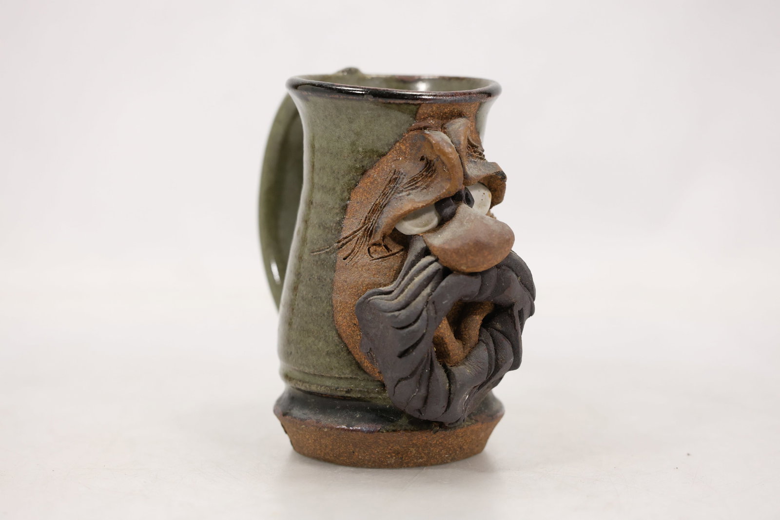 Ryan Signed Studio Pottery Bearded Face Mug: Studio pottery mug with an olive green glaze a cylindrical wheel formed body and a rounded loop handle. The front has an applied sculptural face with modeled eyebrows eyes a projecting nose and a laye