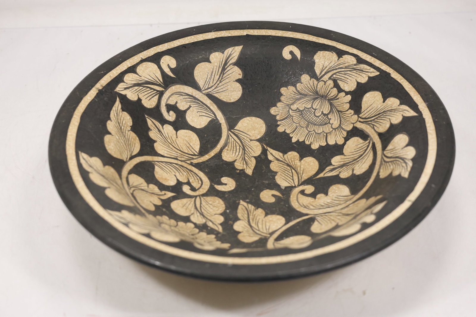 Large Black and Tan Floral Pottery Bowl: Large pottery bowl with wide shallow form tan exterior black rim and black interior decorated with tan floral and scrolling vine design. The bowl has a circular foot ring and rounded sloping sides. Th