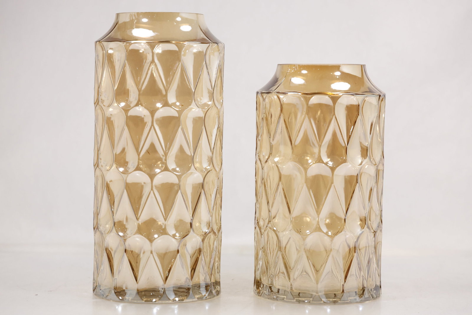 Pair of Amber Pressed Glass Cylinder Vases: Pair of amber glass vases in two graduated sizes with straight cylindrical bodies, short angled shoulders, and flat open rims. Each vase has a molded raised geometric teardrop or diamond pattern aroun