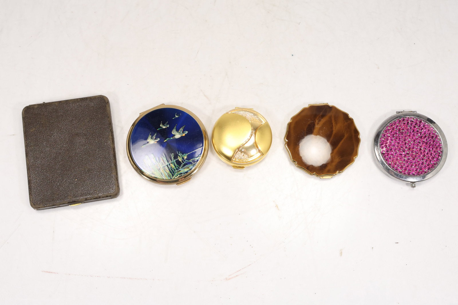 Vintage Powder Compact Group of Five Including Stratton: Group of five powder compacts in varied shapes and finishes. The selection includes a blue enamel round compact with birds and reeds a gold tone modernist compact a brown agate effect compact a rectan