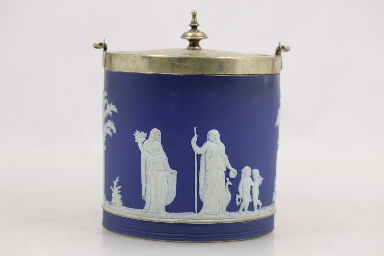 Wedgwood Blue Jasperware Biscuit Barrel with Metal Lid: Wedgwood blue jasperware biscuit barrel with applied white classical relief decoration. The cylindrical vessel is fitted with a metal rim swing handle domed lid and pointed finial. Relief scenes show