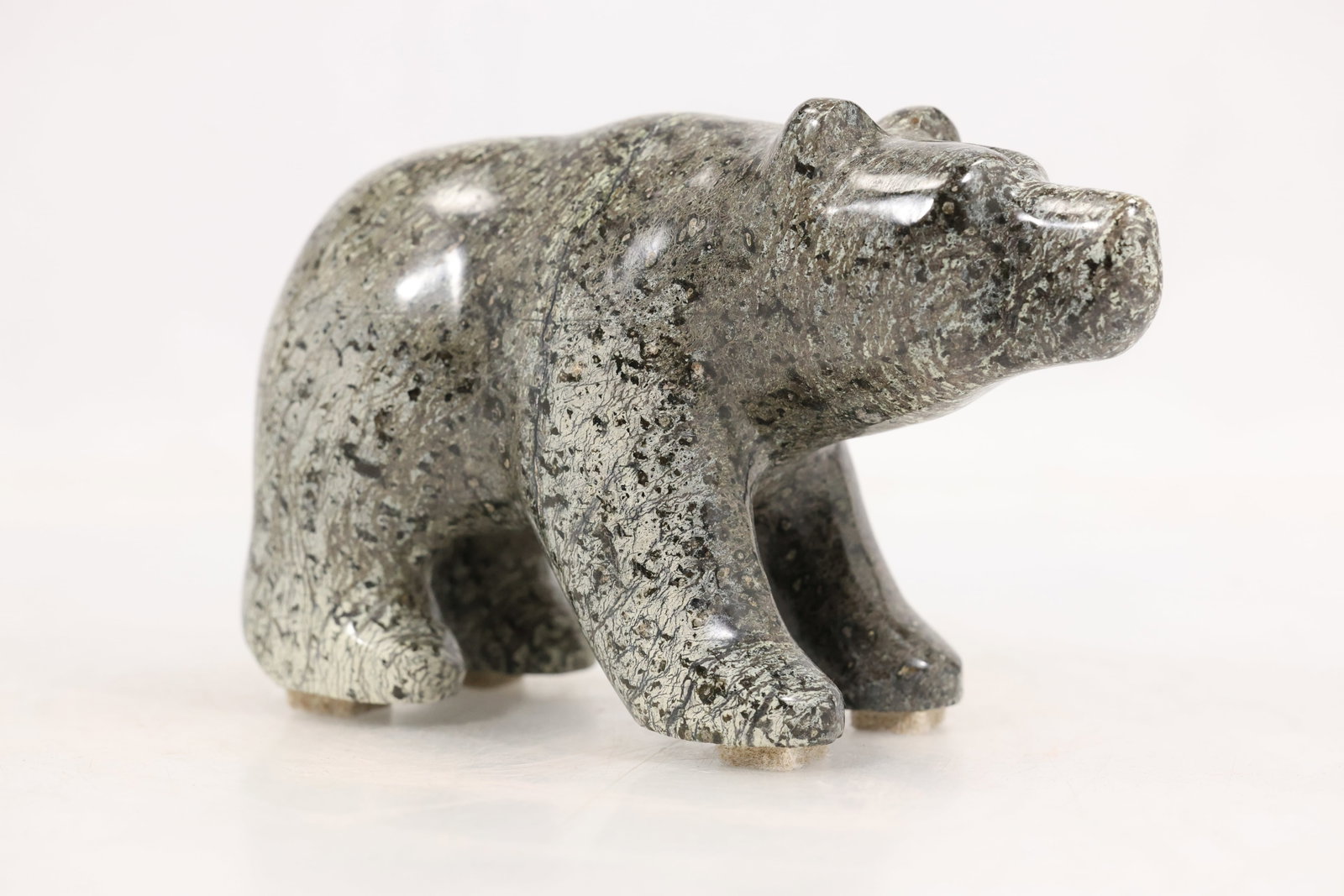 Signed Carved Gray Stone Bear Sculpture: Carved gray stone sculpture of a walking bear with rounded back forward head and simplified block form legs. The polished surface contains dense black and silver colored natural inclusions. An incised