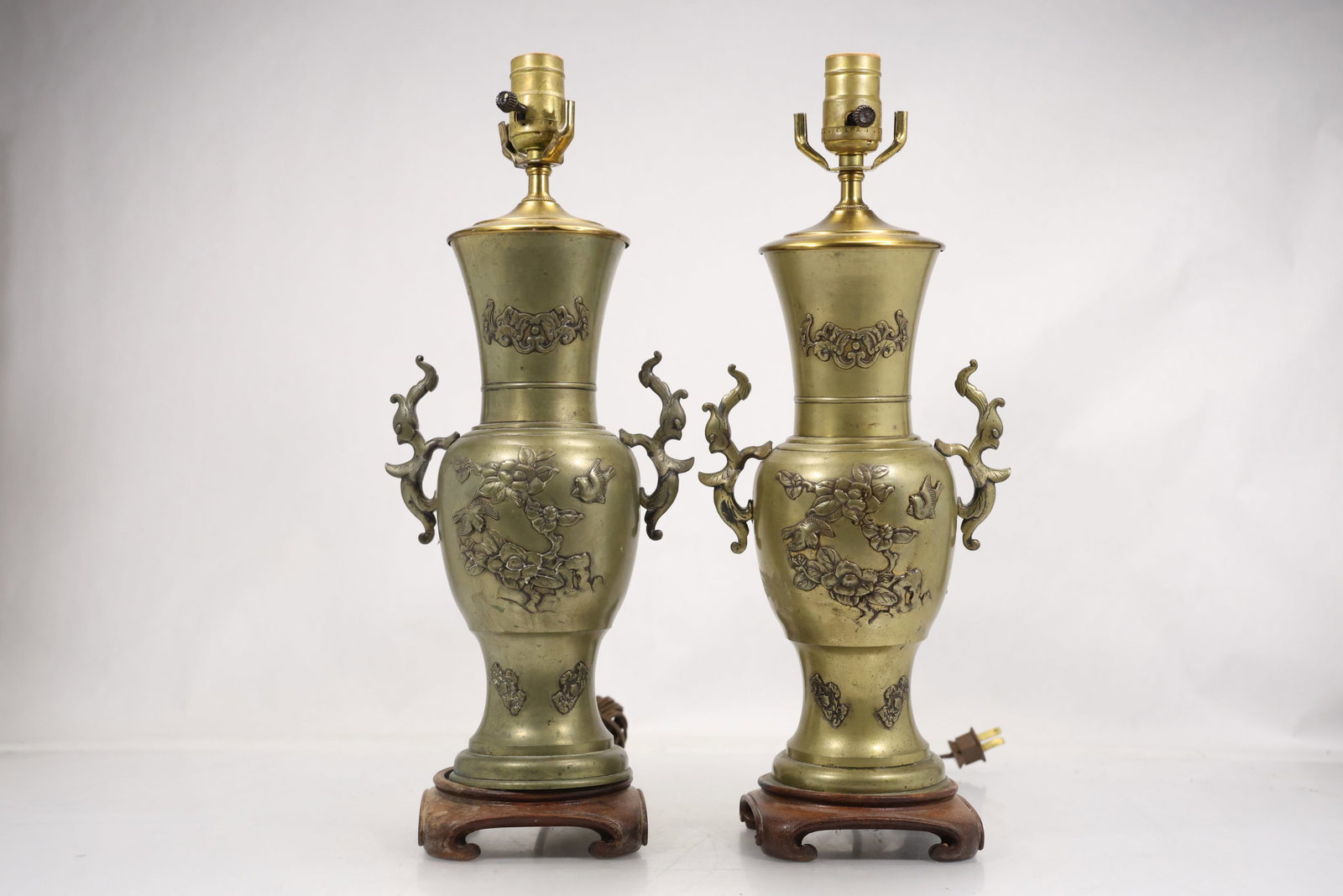 Pair of Chinese Style Relief Decorated Brass Vase Table Lamps: Pair of tall table lamps made from Asian style brass vase forms mounted on carved wood stands. The bodies feature raised landscape bird and floral decoration with archaic scroll and dragon form