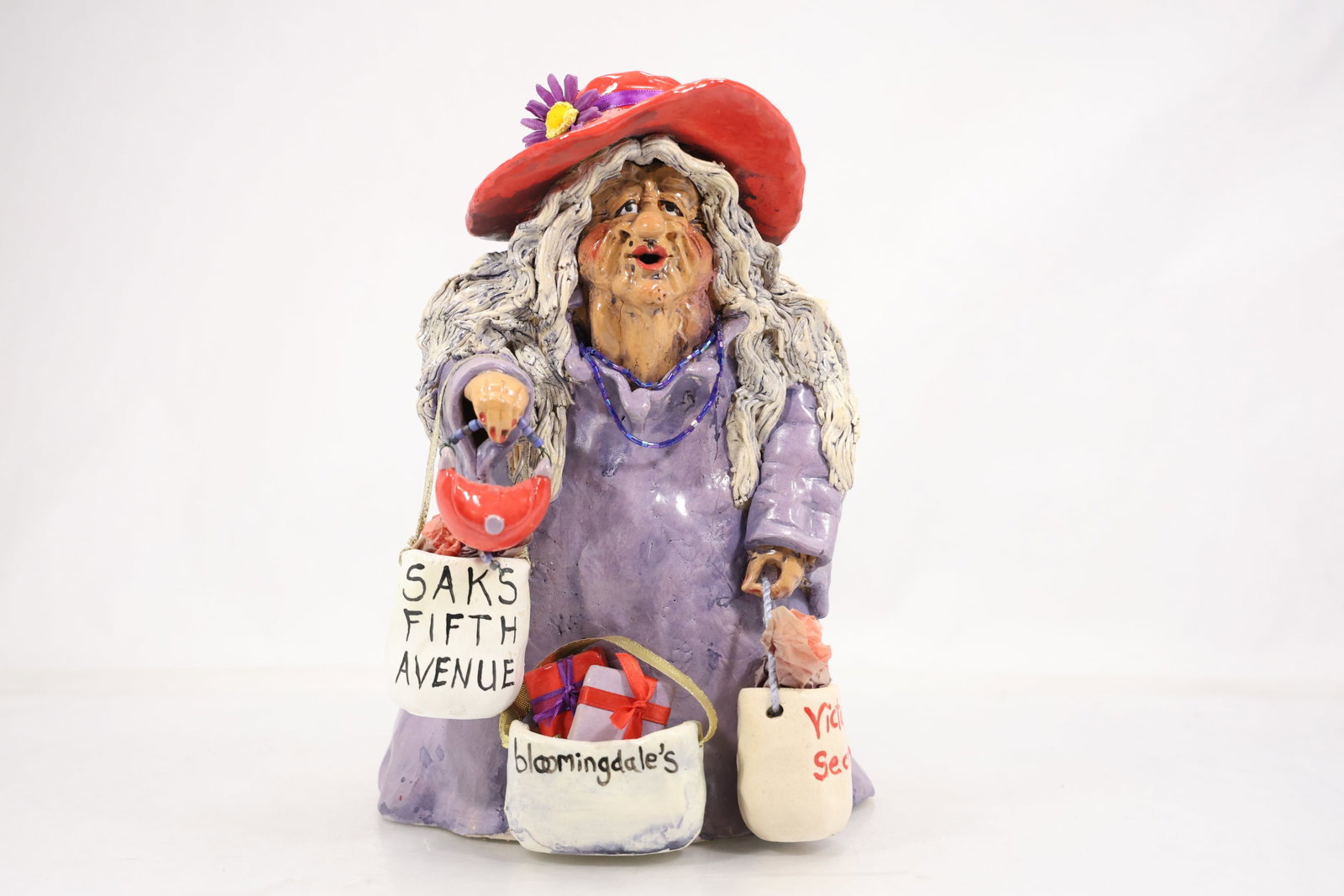 Hand Built Ceramic Shopping Lady Figurine with Store Bags: Hand built and glazed ceramic figure depicting an elderly woman carrying multiple shopping bags. The figure wears a lavender coat and broad red hat with applied flowers. Bags are lettered for Saks