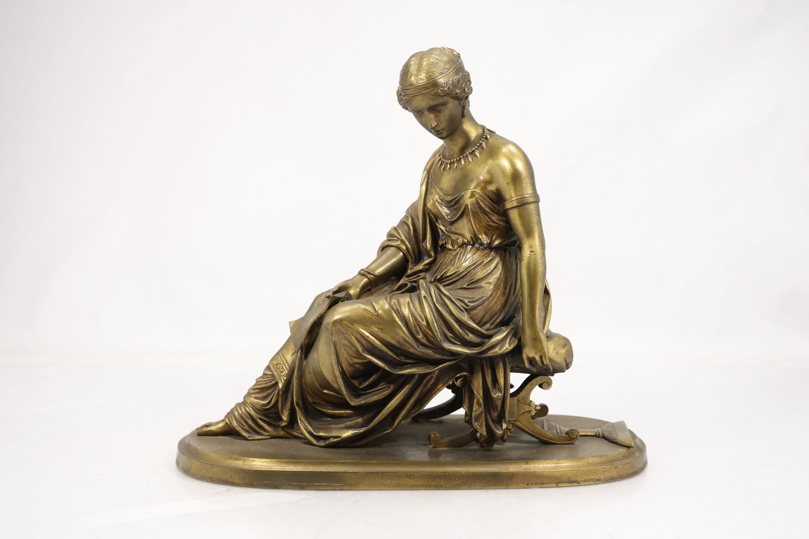 Bronze Tone Seated Classical Woman Sculpture: Bronze tone sculpture of a seated classical woman arranged on an oval base with draped garments and modeled facial features. The figure is presented as a freestanding decorative work. The classical