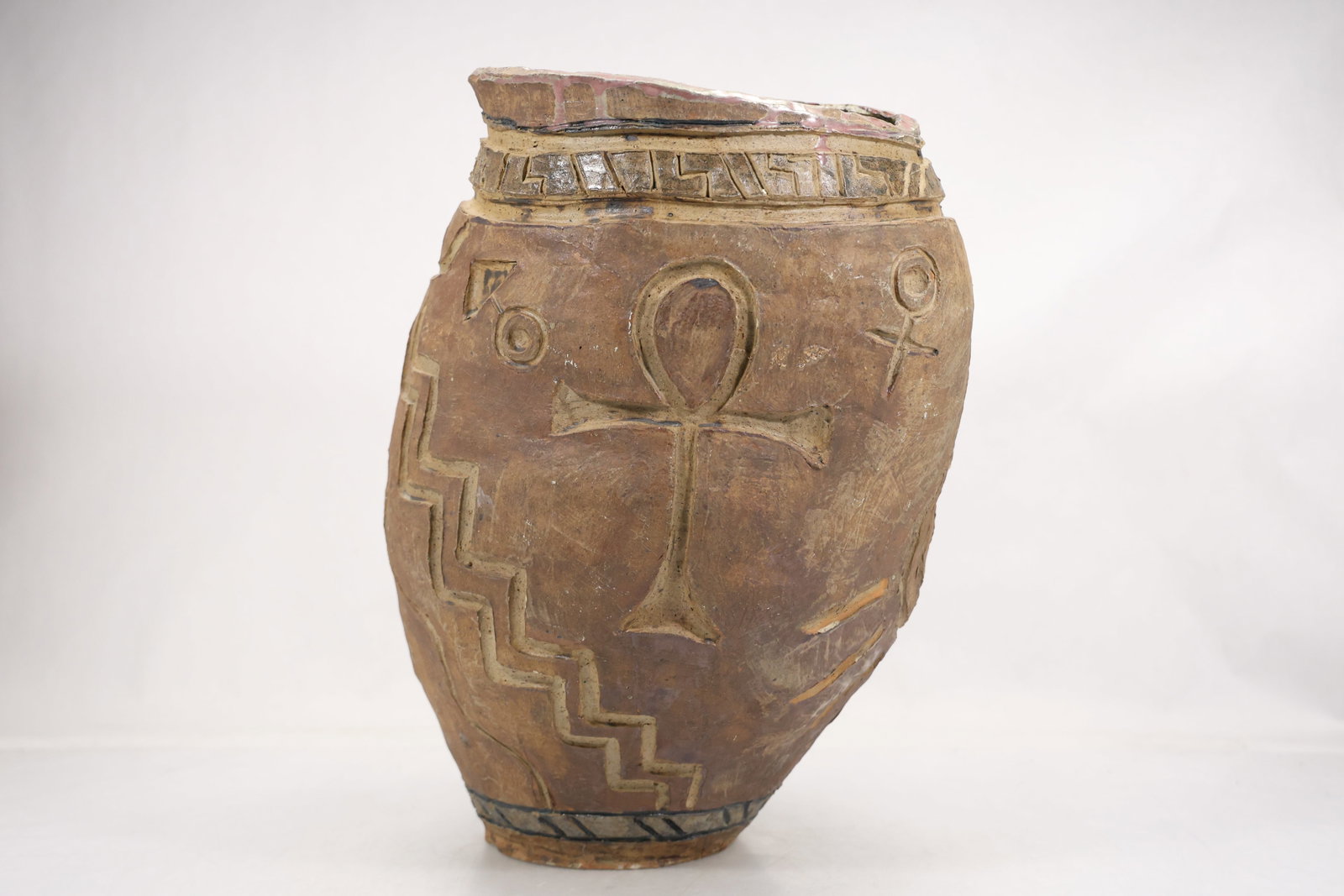 Large Studio Pottery Face and Sun Relief Vase: Large hand built studio pottery vase in warm brown earthen tones with applied relief faces sun motifs and textured geometric passages. The vessel has a broad rounded body and a narrow opening. The