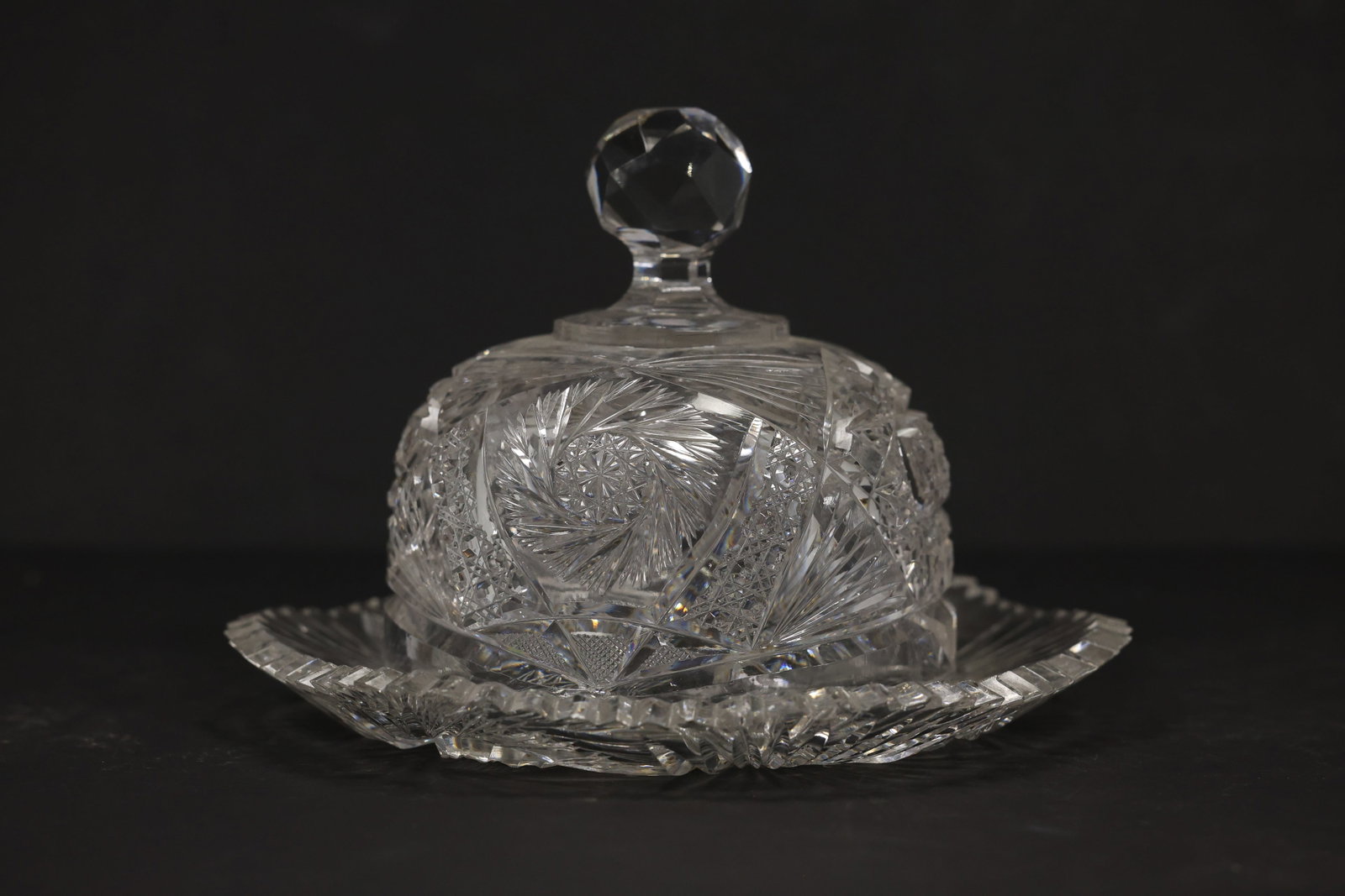 Cut Crystal Covered Butter Dish with Tray: Clear cut crystal covered serving dish with a domed lid faceted ball finial and matching oval tray. Starburst fan and geometric cuts cover the coordinated pieces. The dense cut pattern relates to Amer