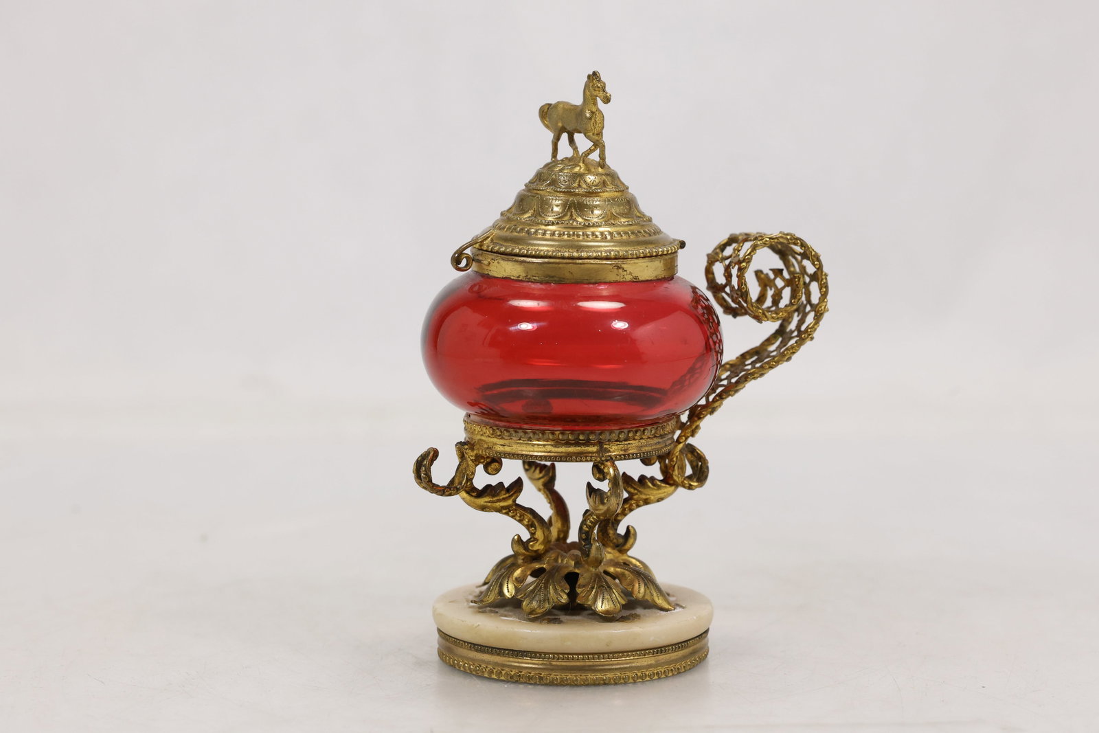 Red Glass and Gilt Metal Horse Finial Vessel: Decorative red glass vessel mounted with ornate gilt tone metal bands handles and a domed cover topped by a horse finial. The piece rests on a dark stone form base. The combination of colored glass