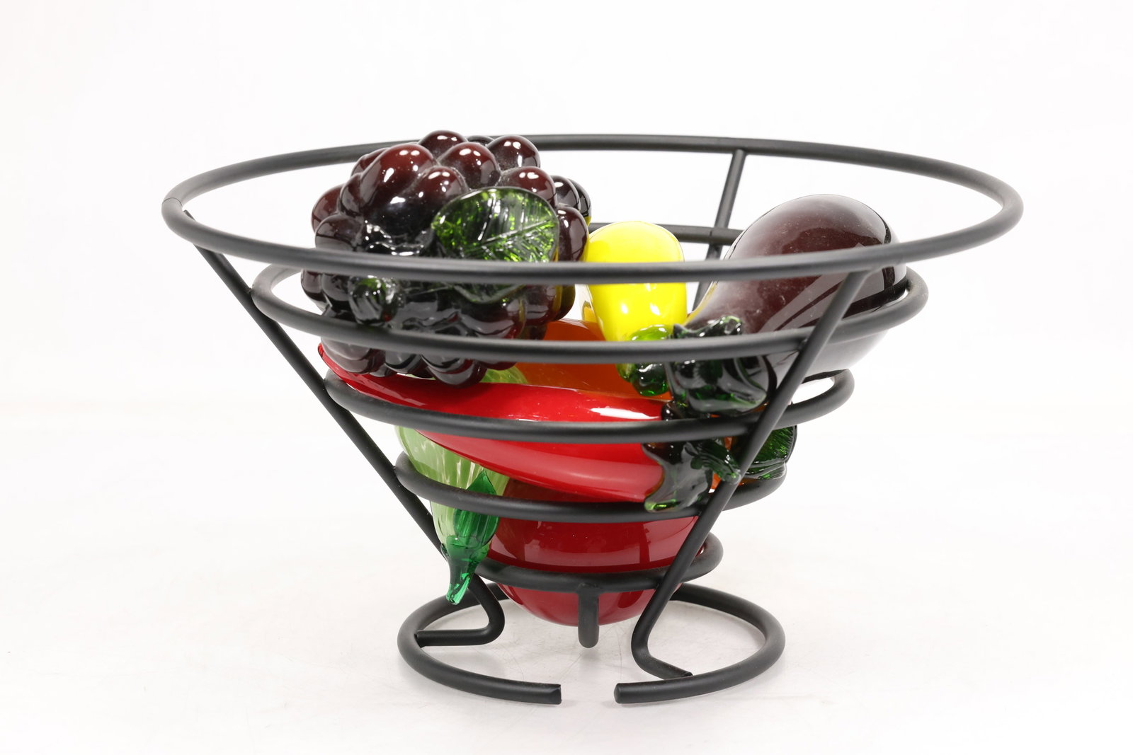Nine Art Glass Fruits with Metal Basket: Group of nine colorful art glass fruits displayed in a black metal openwork basket. The assortment includes grapes banana pear peppers and other molded fruit forms in red yellow green and purple. The