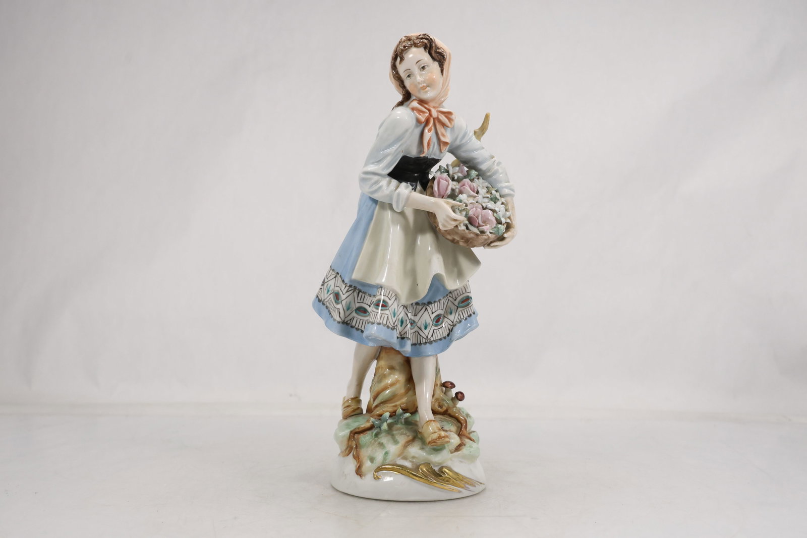 Porcelain Flower Seller Woman Figurine: Porcelain figurine of a young woman in a head scarf apron and blue skirt holding a basket filled with modeled flowers. The figure stands beside a tree form support on a gilt accented naturalistic base