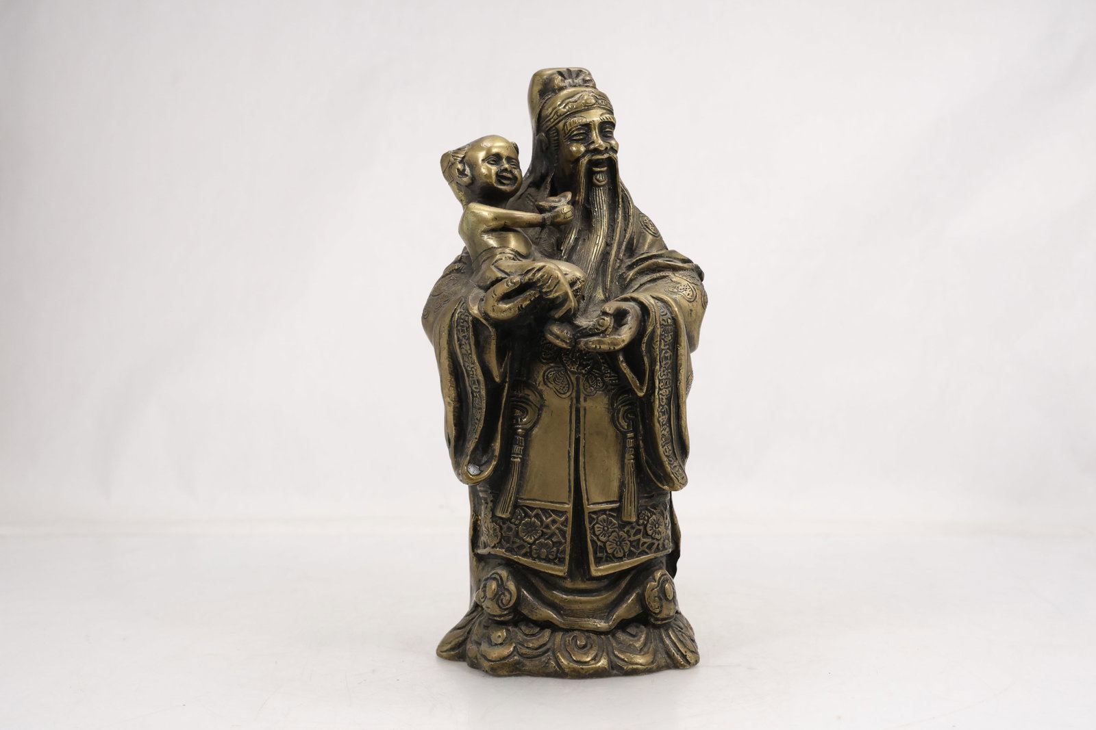Chinese Brass Tone Elder and Child Figure: Cast brass tone Chinese figure depicting a bearded robed elder holding a small child. Incised floral and geometric patterns decorate the garments and lotus form base. The subject relates to Chinese re