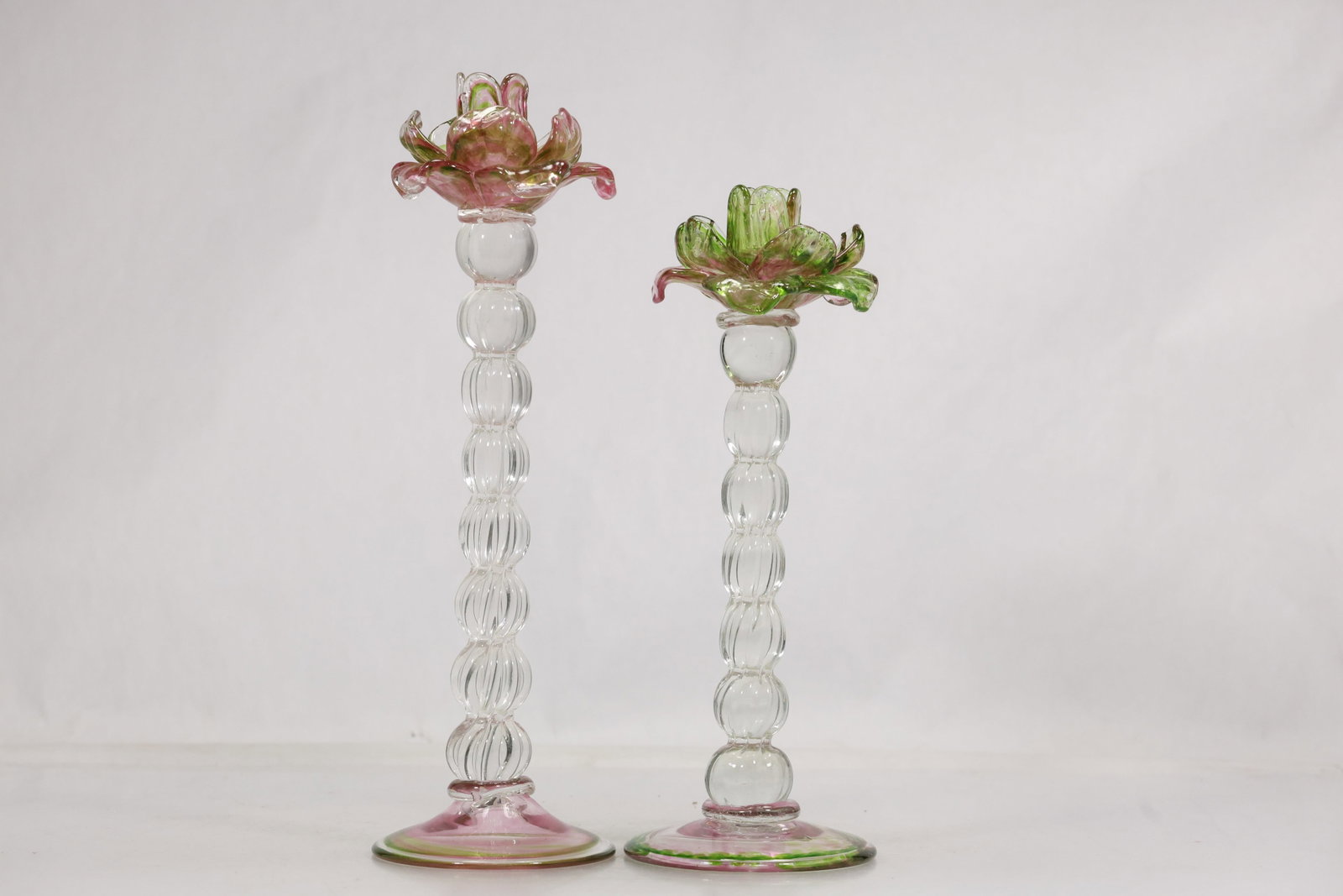 Pair Tall Glass Flower Candlesticks: Pair of tall clear glass candlesticks with stacked ribbed ball stems and applied flower form candle cups. One is accented in pink and the other in green with coordinating circular feet. The floral fan
