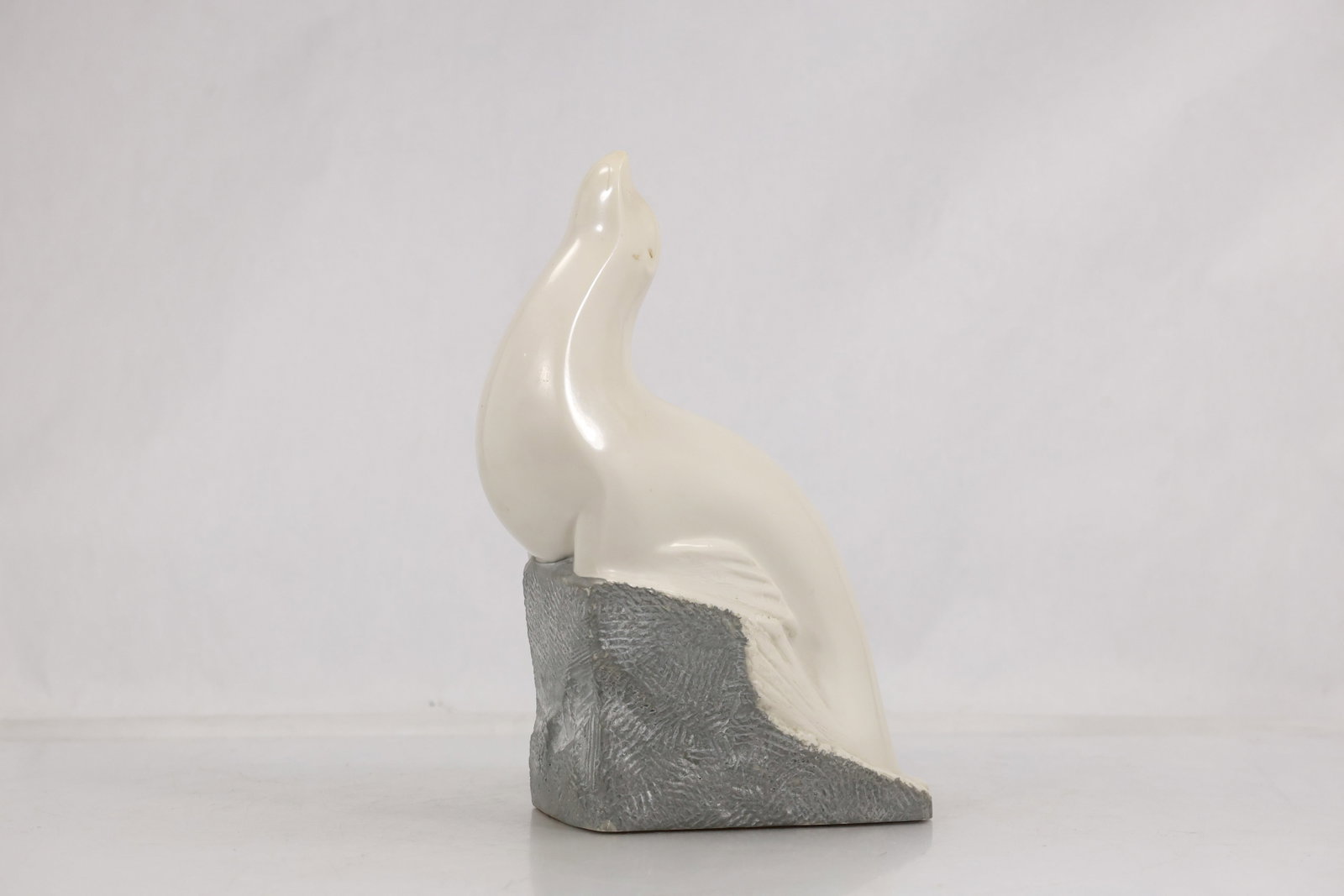 V Glinsky Carved Stone Seal Sculpture on Textured Base: Carved stone sculpture depicting a stylized seal in an upright pose atop an angular textured base. The figure features a smooth polished cream colored surface contrasted by a textured gray base that e