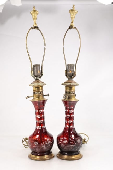 Pair Bohemian Style Ruby Cut to Clear Glass Table Lamps: Matched pair of table lamps with ruby overlay glass bodies cut to reveal clear oval and floral motifs. Each lamp has a slender neck rounded lower body etched foliate band brass tone circular base burn