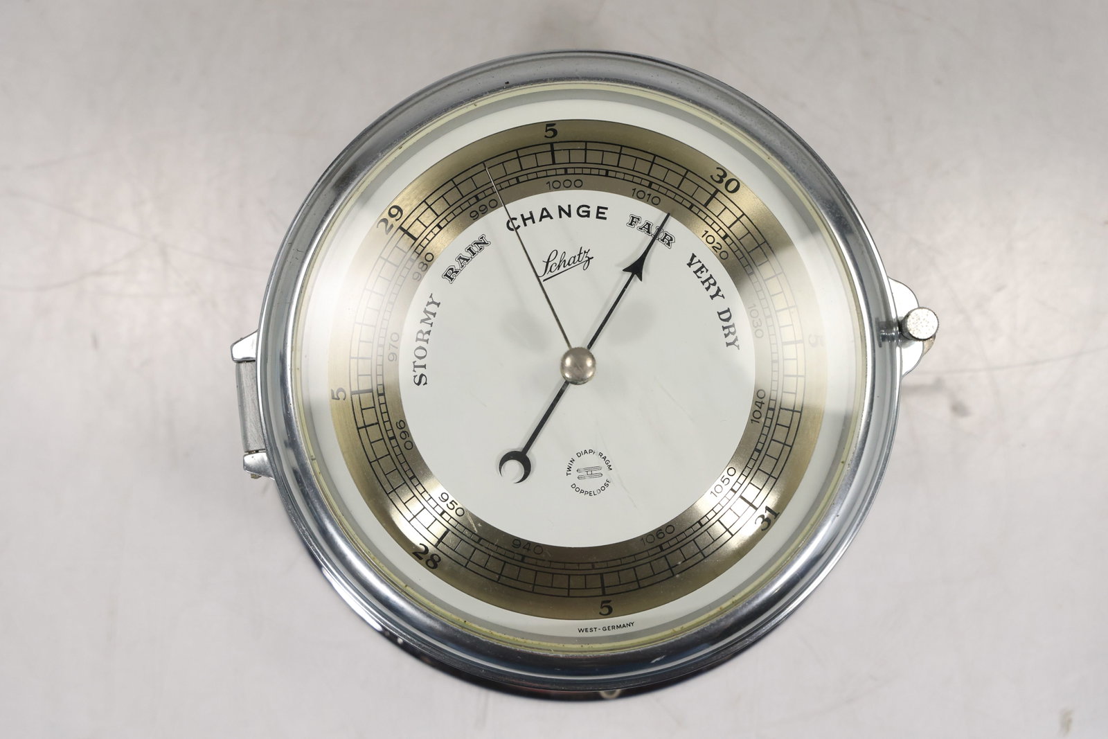 Schatz German Chrome Aneroid Barometer with Hinged Glass Bezel: Schatz aneroid barometer in a polished chrome tone cylindrical case with a flared circular base and hinged glass bezel. The white and metallic dial displays atmospheric pressure scales and the weather