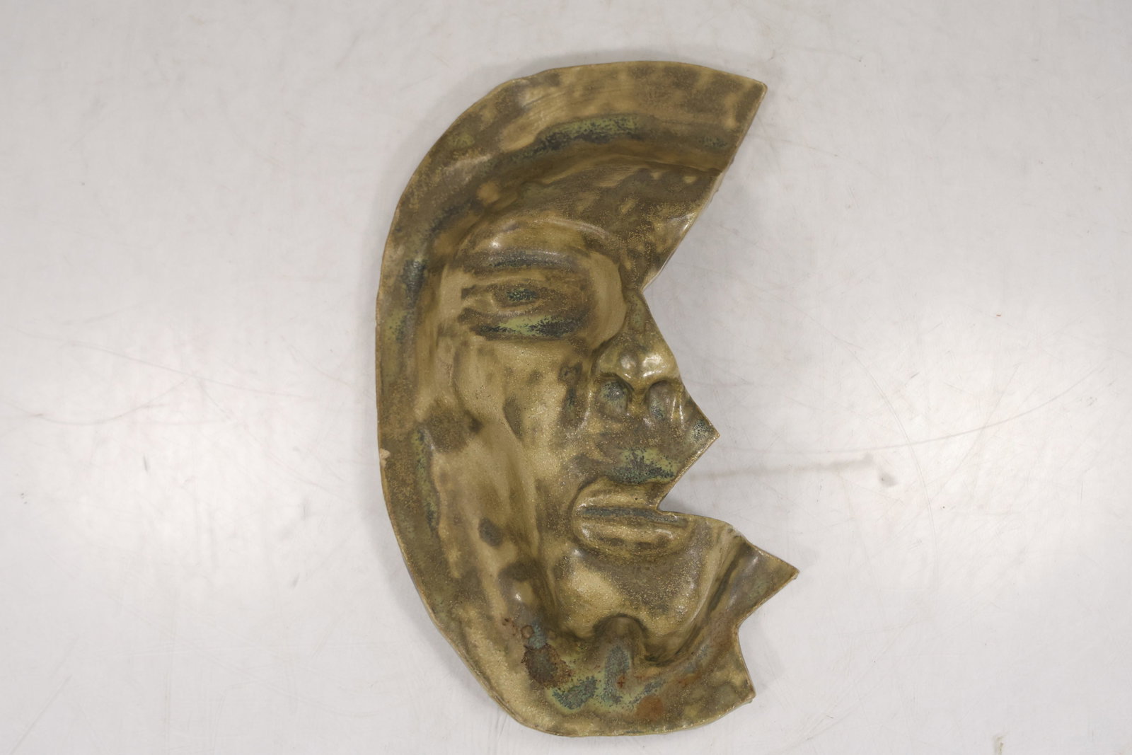 Abstract Ceramic Face Mask Sculpture: Abstract ceramic or pottery face sculpture with crescent shaped form raised facial features and mottled tan brown green and dark glaze. The piece has a partial face profile with modeled eye nose mouth