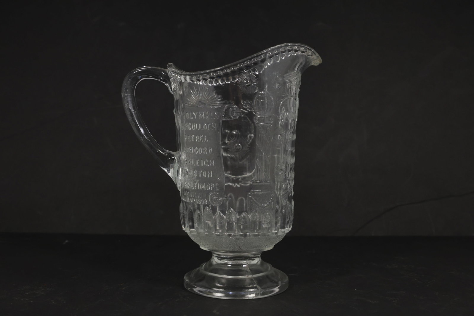 Admiral Dewey Manila Bay Pressed Glass Pitcher: Clear pressed glass commemorative pitcher with handle, pedestal foot, beaded rim, spout, and molded Spanish American War imagery. The molded decoration includes an eagle with shield, naval and