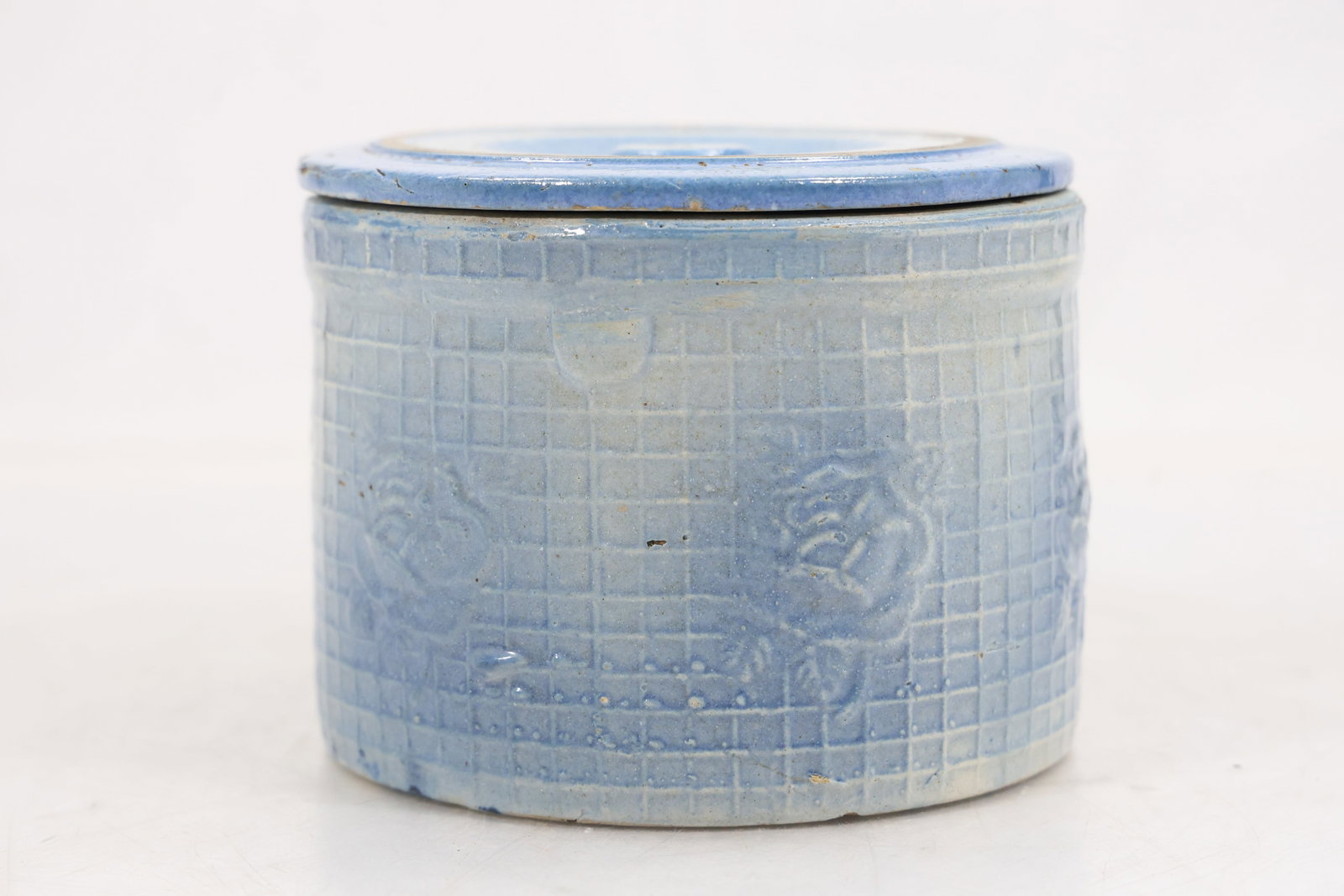 Blue Glazed Stoneware Lidded Crock with Lion Medallions: Cylindrical pale blue glazed stoneware crock with fitted circular lid and knob. The body has a textured grid ground with repeating raised crest and lion medallions. The base appears unmarked and the