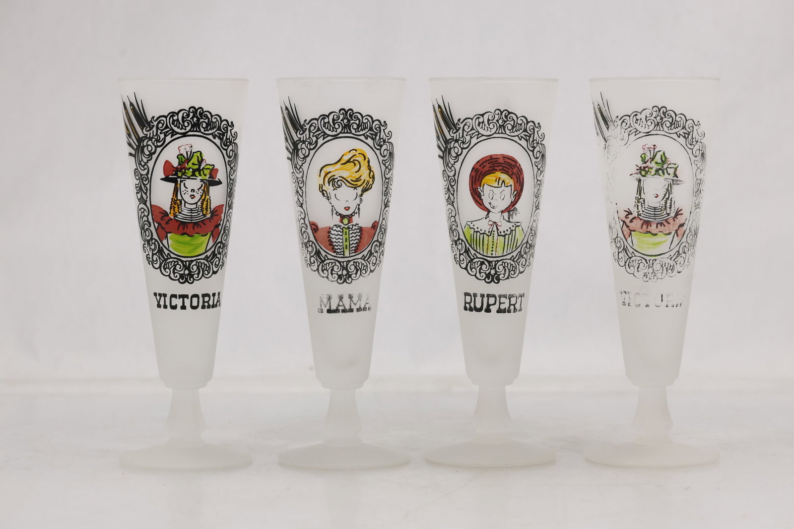 Four Frosted Character Portrait Pilsner Glasses: Set of four tall frosted glass pilsner vessels decorated with illustrated character portraits and printed names including Victoria Mama and Rupert. Each glass has a flared foot and a clouded white fin