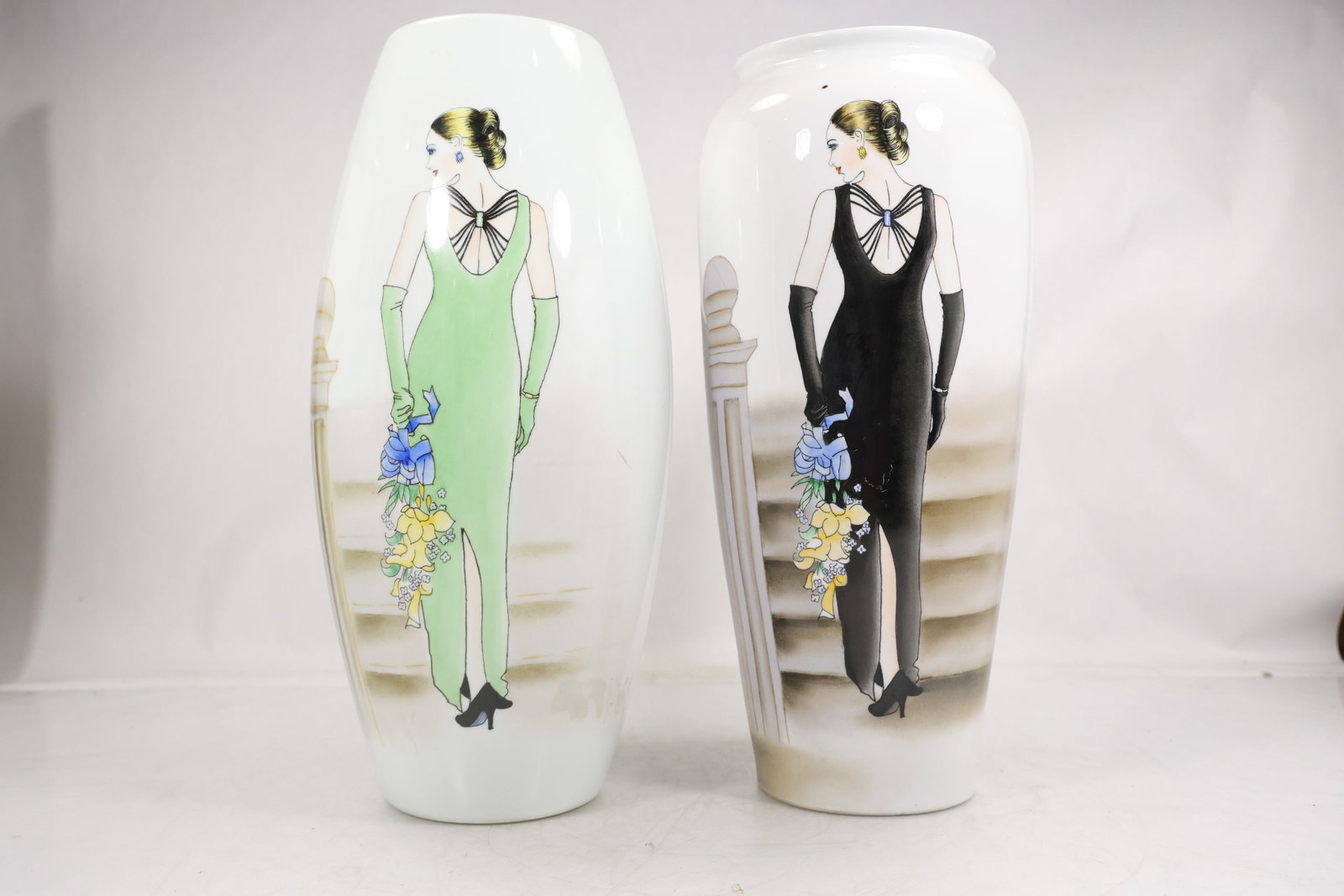 Pair of Tall Art Deco Style Porcelain Vases – Flapper Lady Design: Matching pair of tall porcelain vases featuring elegant Art Deco-style flapper figures. Each vase depicts a stylized woman in period dress—one in soft green, the other in black—holding floral bouq