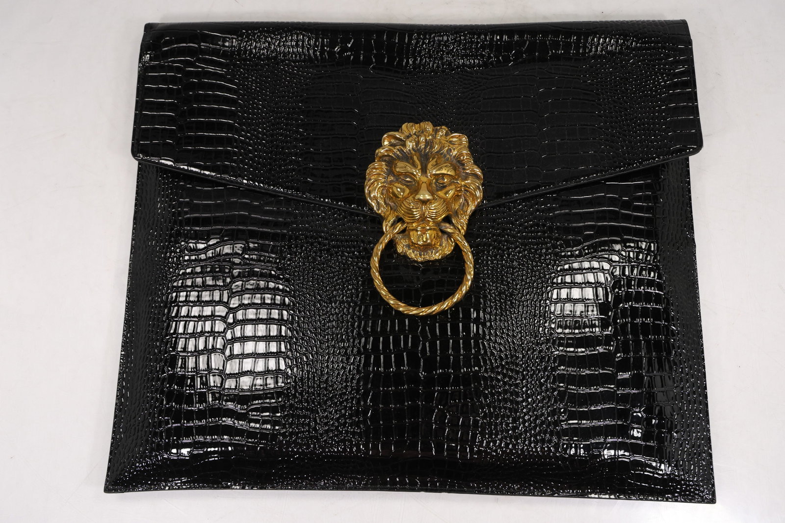 Black Faux Crocodile Clutch Purse with Lion Head Ring Closure: Envelope style clutch purse finished in black faux crocodile embossed material with a glossy surface. The front flap is fitted with a prominent gold tone lion head ornament holding a twisted ring pull